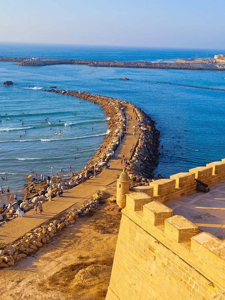 Yellow fortress walls frame a view of beach crowds on a curved jetty leading to calm blue ocean waters