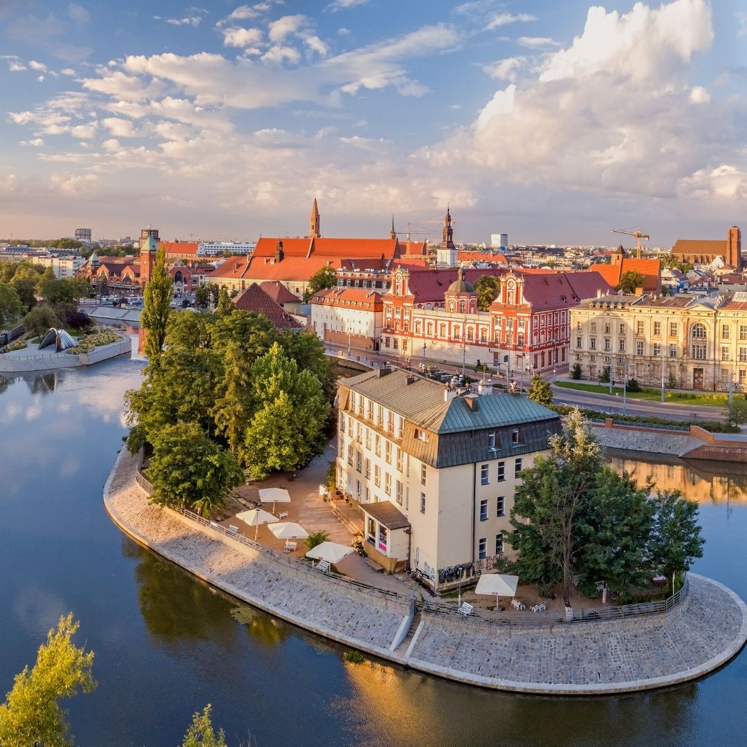 Historic riverside town in Poland with red roofs, trees, and calm water under a cloudy sky