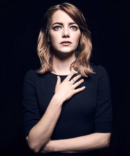 Portrait of Emma Stone in a black top with her hand on her chest against a dark background