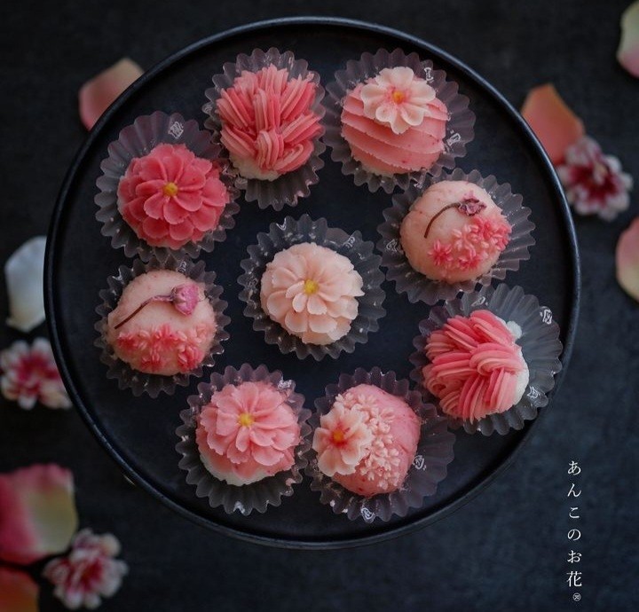 Nine pink flower-shaped Japanese wagashi pastries arranged on a round black plate with scattered petals