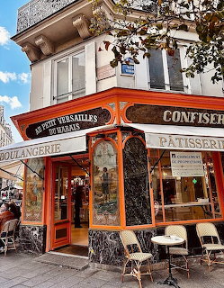 Red-fronted corner bakery with ornate awnings and outdoor cafe tables on sidewalk