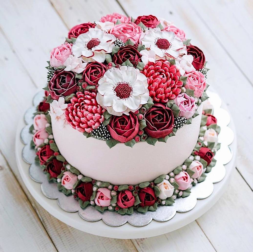 Two-tier white cake adorned with pink, red, white, and burgundy buttercream flowers on wooden background