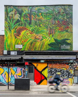 Large tiger mural on a city building above graffiti-covered storefronts, with a cyclist riding past