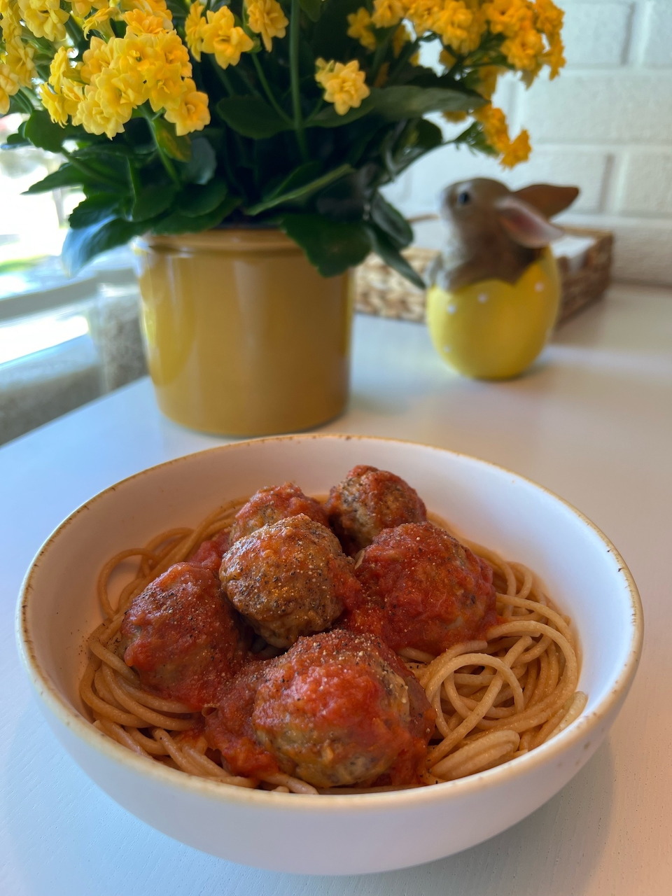 White bowl of spaghetti and meatballs in tomato sauce on a counter with yellow flowers behind