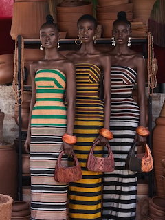 Three African models wearing colorful striped strapless dresses holding brown handbags near clay pottery