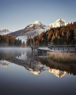 Calm alpine lake reflecting snowy mountains and orange autumn trees with a wooden pier in morning mist