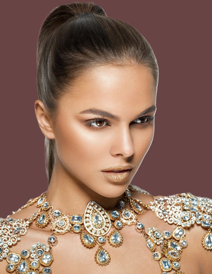 Woman with slicked-back ponytail wearing ornate gold and crystal necklace against brown background