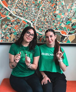 Two women in green shirts posing in front of a colorful abstract map artwork for St. Patrick's Day