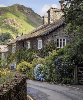 Stone cottage with chimneys beneath a green hill with blue hydrangeas and dry stone wall in Yorkshire