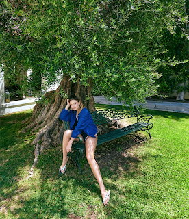 Woman in blue dress sitting on bench beneath tree in sunny garden