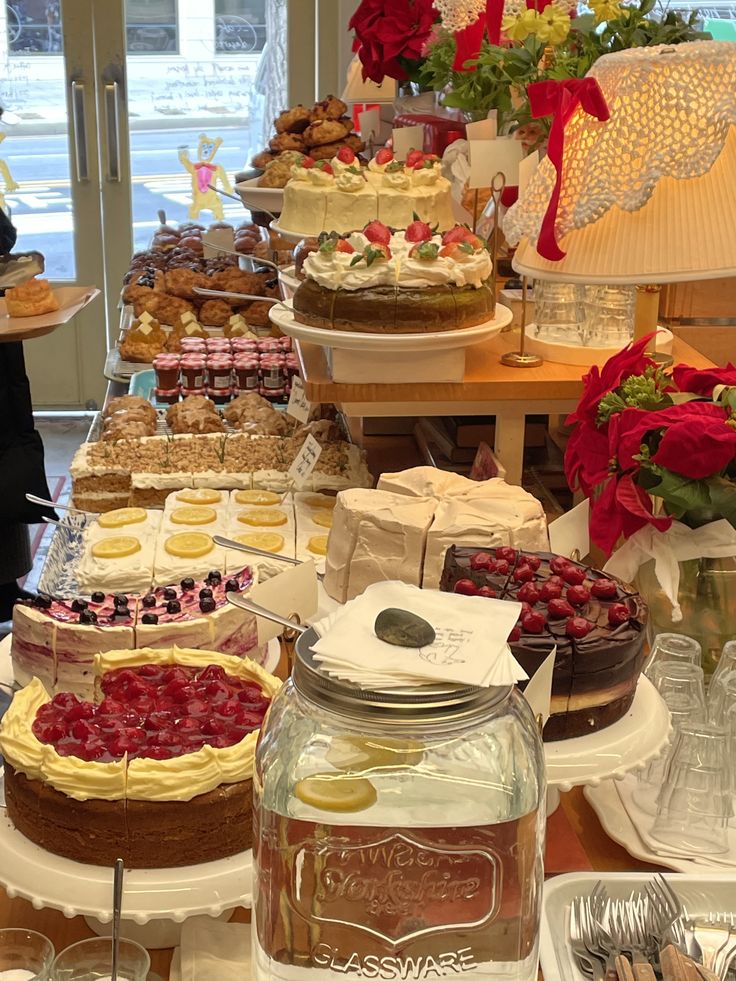 Cafe buffet table filled with assorted cakes, berry tarts, pastries, and a glass jar under warm lamp light