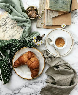 Croissant on plate beside espresso cup, green linen, daisies and old book on marble