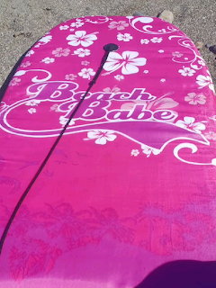 Pink surfboard with white hibiscus floral pattern and Beach Babe letering on sand
