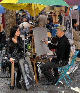 Street artist drawing a portrait of a seated woman at an outdoor art market with umbrellas