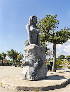 Large grey mermaid statue sitting on a rock in a town square with trees and blue sky