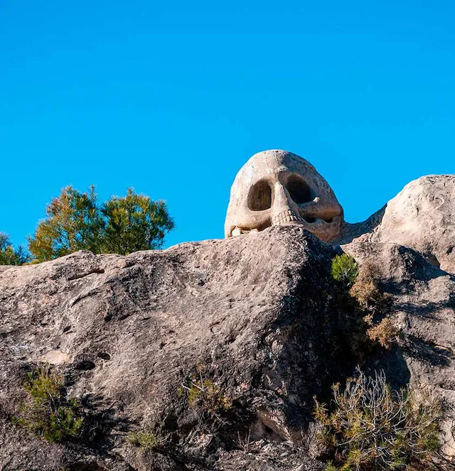 Skull-shaped stone carving on a rocky hillside beneath a clear blue sky
