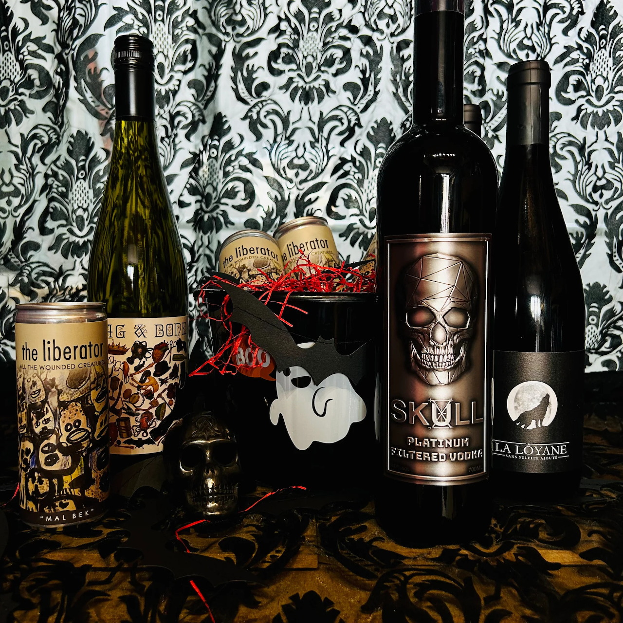 Four wine bottles with Halloween skull and gothic labels arranged on table with decorative pumpkin glass and damask backdrop