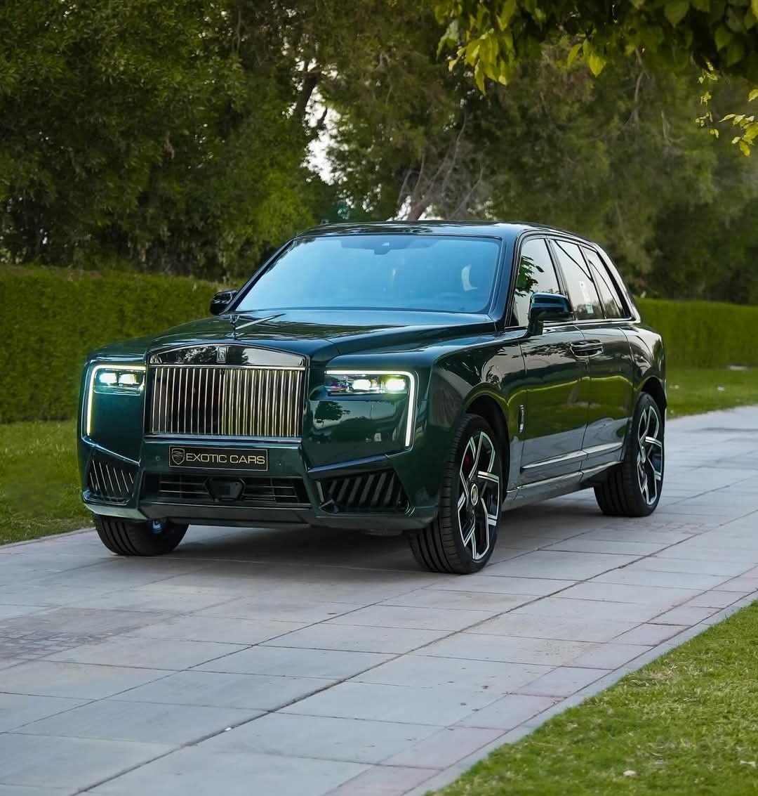 Dark green Rolls-Royce Cullinan SUV on a paved driveway with trees and lawn behind it.