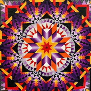 Colorful geometric quilt with starburst center and symmetrical triangular border patterns in purple, orange, yellow, red