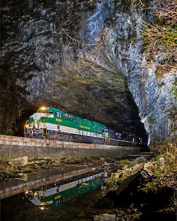 Green freight train in a rocky natural tunnel above reflective water