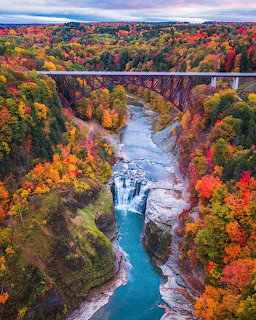 Aerial view of autumn gorge with waterfall, turquoise river, colorful trees, and railway bridge crossing canyon