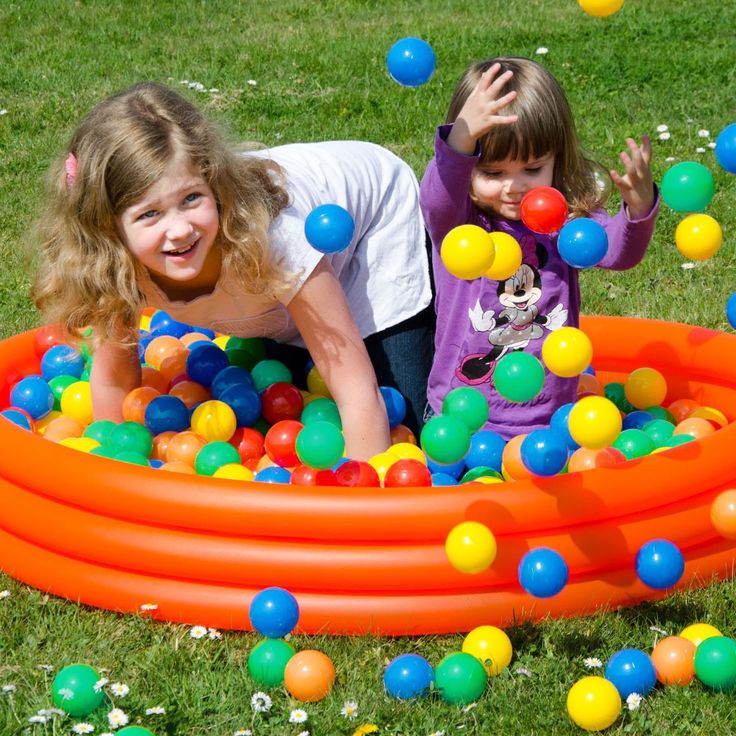 Two girls playing in an orange inflatable pool filled with colorful balls on grass
