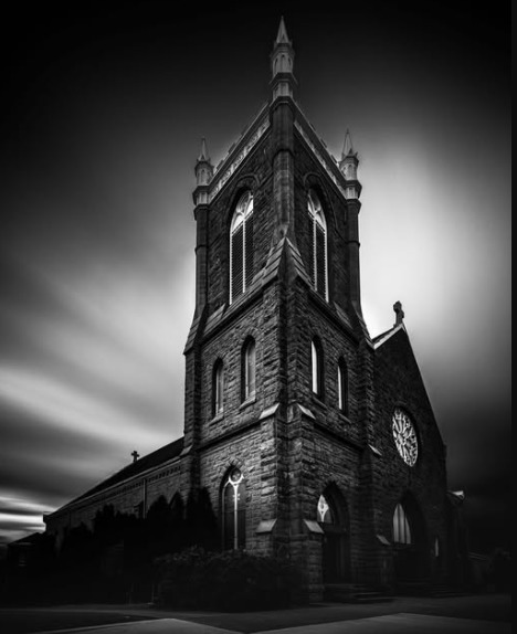 Dramatic black and white photo of a tall Gothic stone church with streaking clouds overhead