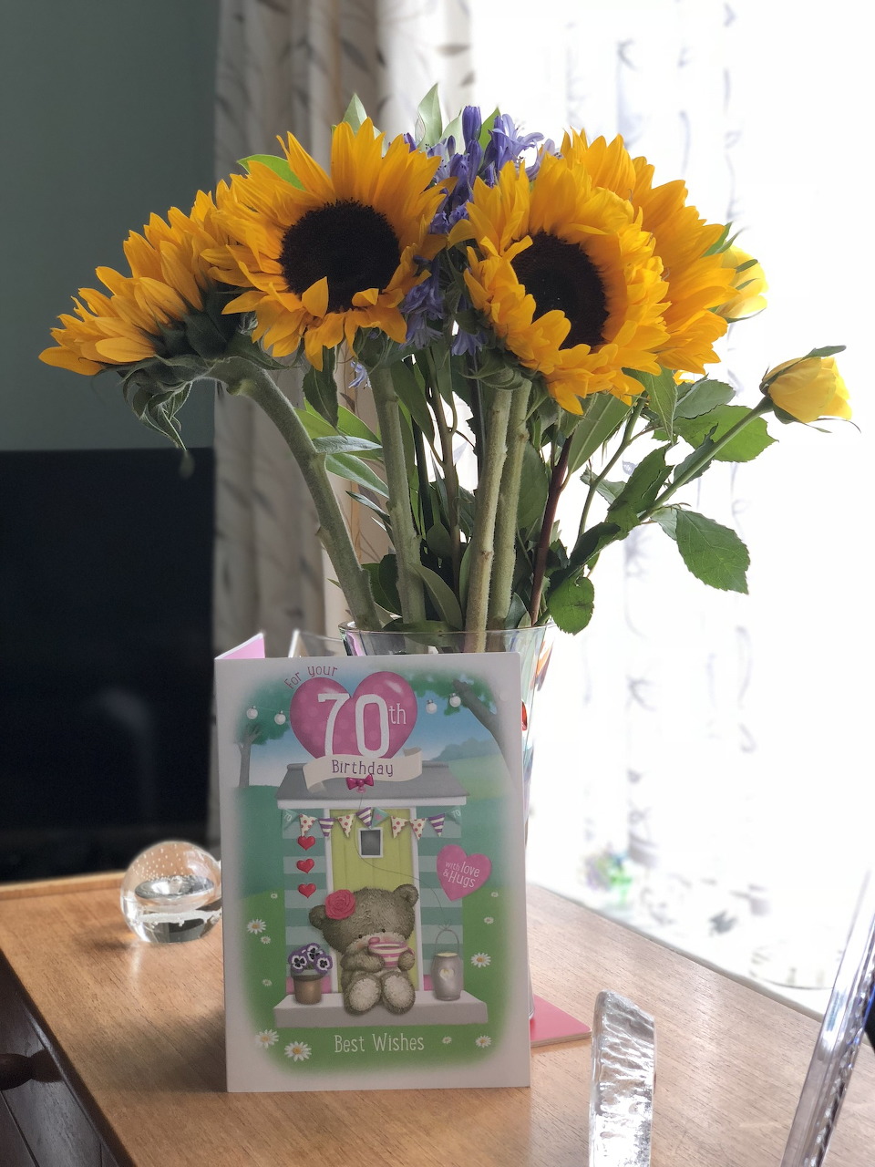 Yellow sunflowers in a white vase next to a 70th birthday card on a wooden table
