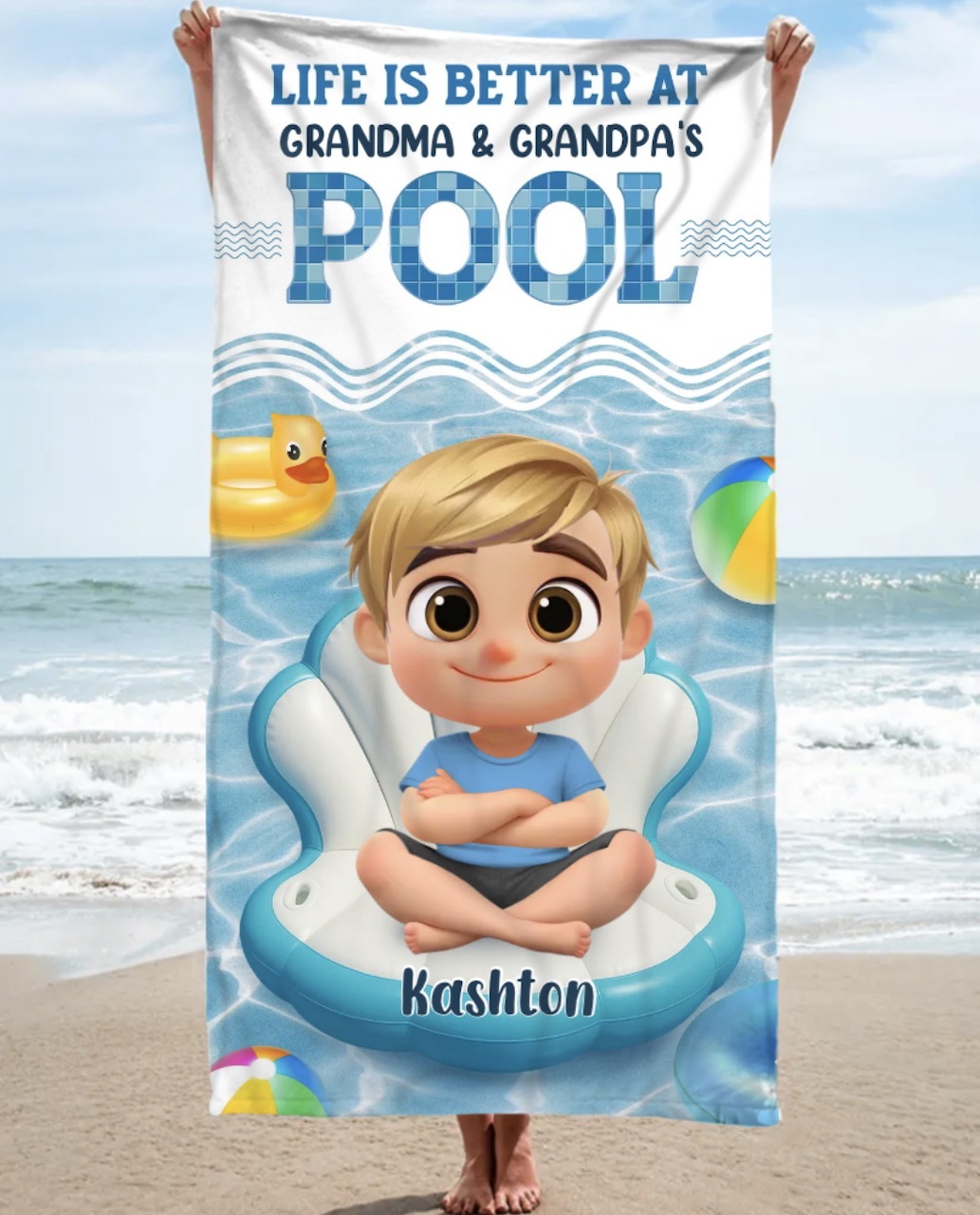 Beach towel with a cartoon boy in a float and pool-themed text, held in front of the ocean