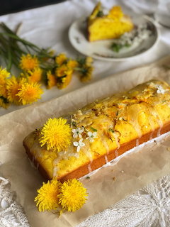 Glazed loaf cake on parchment with yellow dandelions and a blurred slice on a plate behind