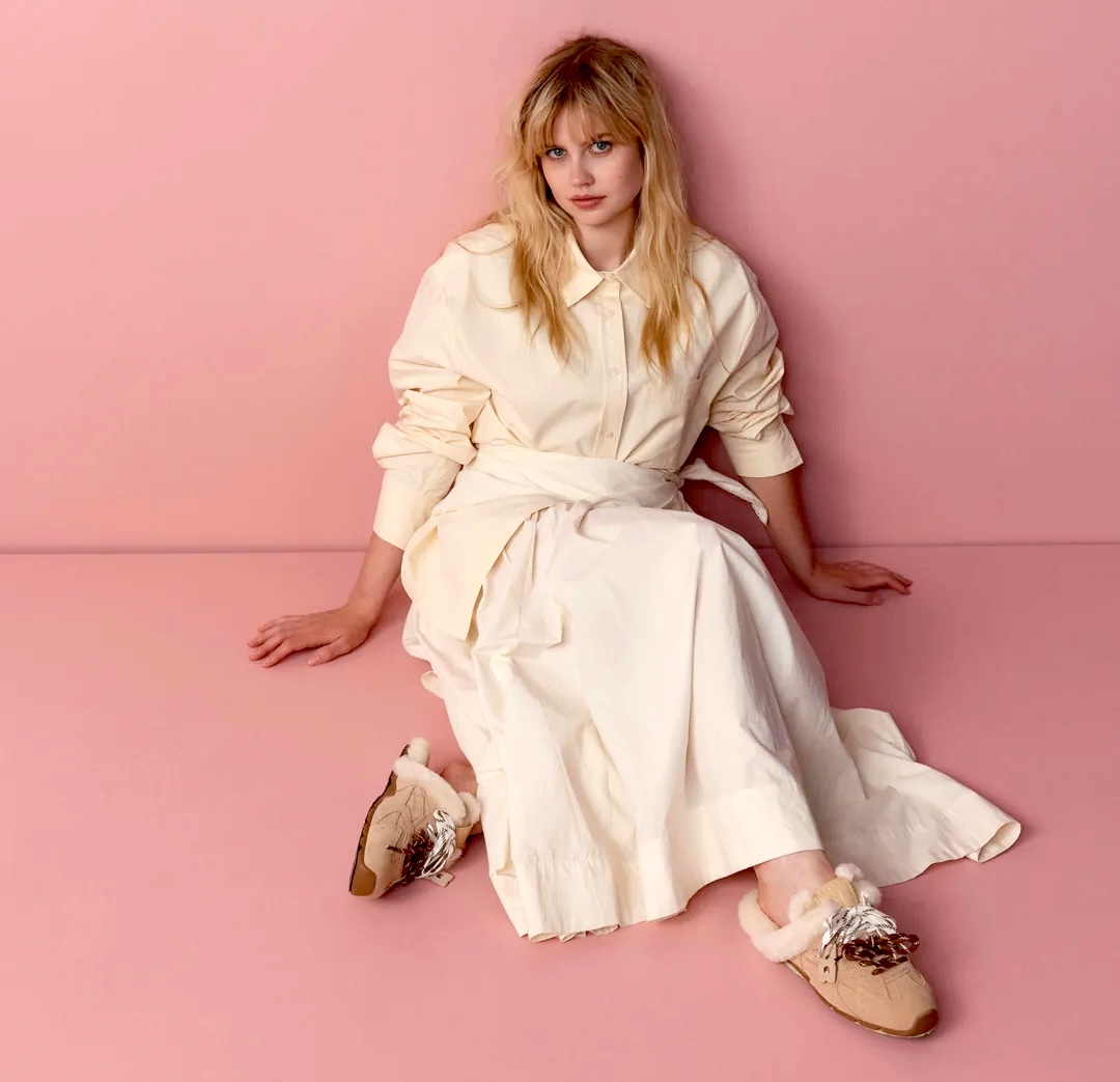 Blonde woman in white dress sitting on pink floor against pink background