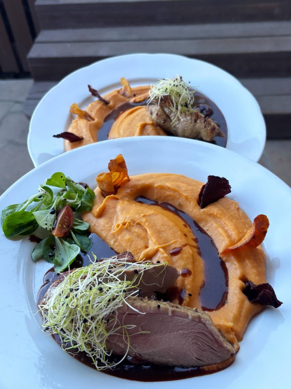 Two white plates with sliced duck breast, sweet potato chips, microgreens, and dark sauce on outdoor table