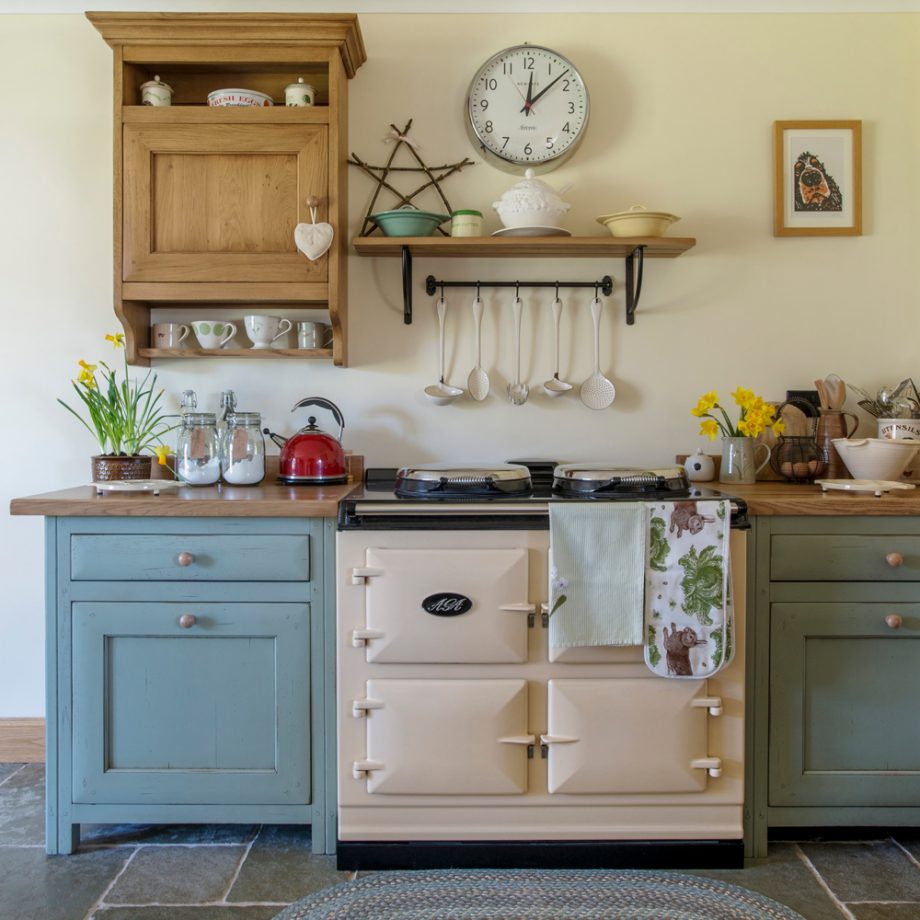 Cozy country kitchen with cream stove, blue cabinets, wooden shelves, clock, and flowers on countertop