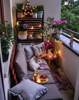 Cozy balcony with cushions, a small dog, potted pink flowers, candles, and string lights at dusk