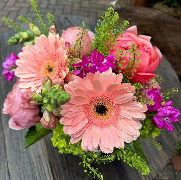 Vibrant bouquet of pink gerbera daisies, roses, and purple flowers with green foliage on wooden surface