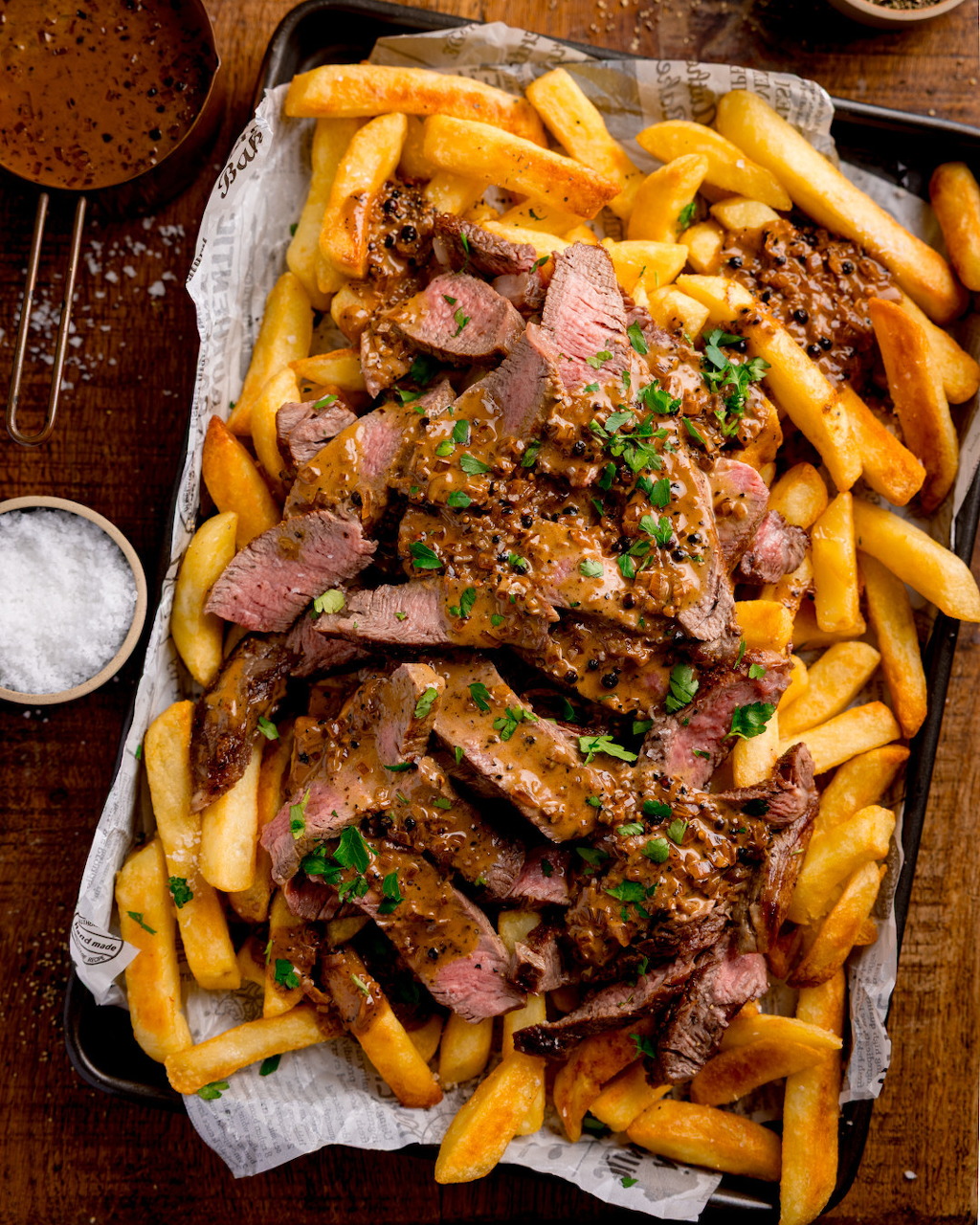 Sliced grilled steak on thick-cut chips in a baking tray, garnished with herbs and sesame seeds