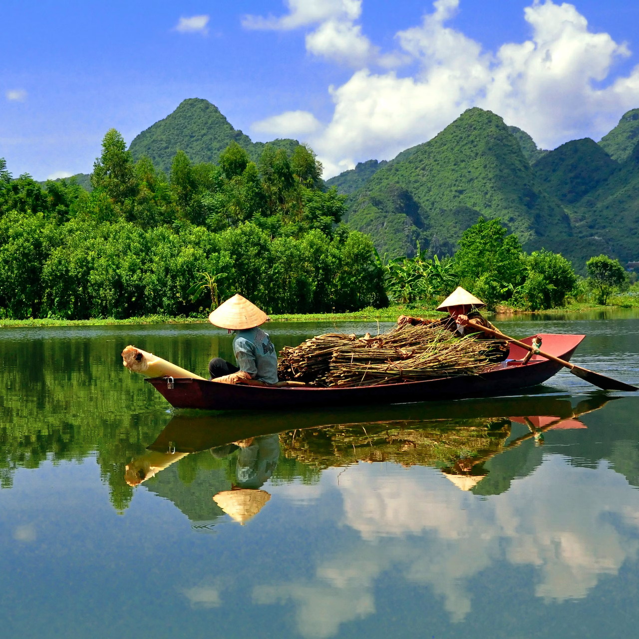 Small boat with two people in conical hats on a calm river with mountain reflections