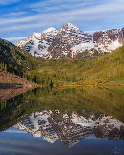 Snow-capped Maroon Bells mountains reflected in a still alpine lake with green forest in foreground