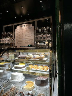 Glass cafe display case with various cakes, pastries, and desserts under warm lighting with menu board above