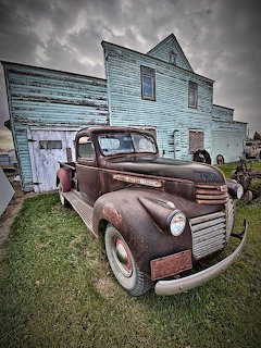 Rusty vintage GMC pickup truck in front of an old blue wooden building under cloudy sky