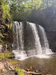 Wide waterfall flowing over rocky ledge into pool surrounded by forest with people viewing from rocky shore