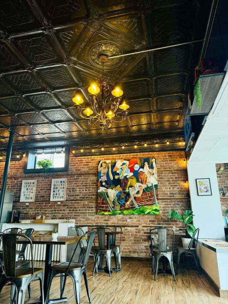 Cafe interior with brick walls, tin ceiling, chandelier, string lights, and colorful mural above dining tables