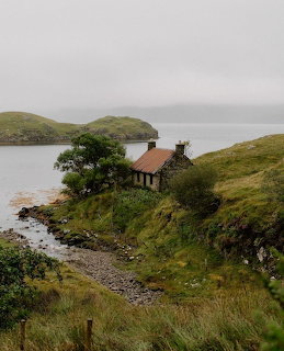 Stone cottage with red roof on hillside beside Scottish loch with overcast sky