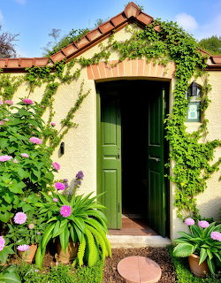 Green wooden door surrounded by lush ivy and purple flowers on cream cottage with red tile roof
