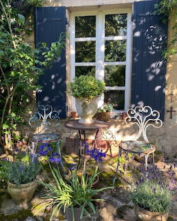Blue-shuttered cottage window with wrought iron table and chair surrounded by poted plants and flowers