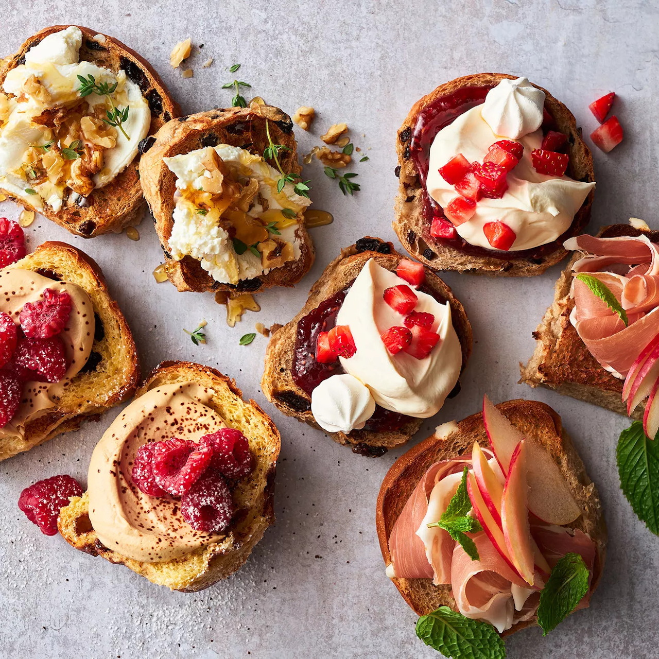 Variety of gourmet toast with strawberries, cream cheese, prosciutto, and fresh herbs arranged on stone background