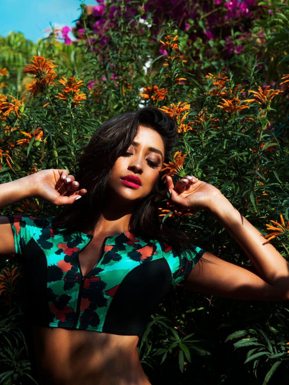 Dark-haired woman posing among orange flowers and greenery wearing a floral crop top