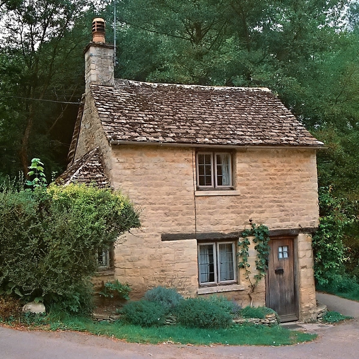 Small historic stone cottage with aged walls, windows, and chimney surrounded by lush greenery