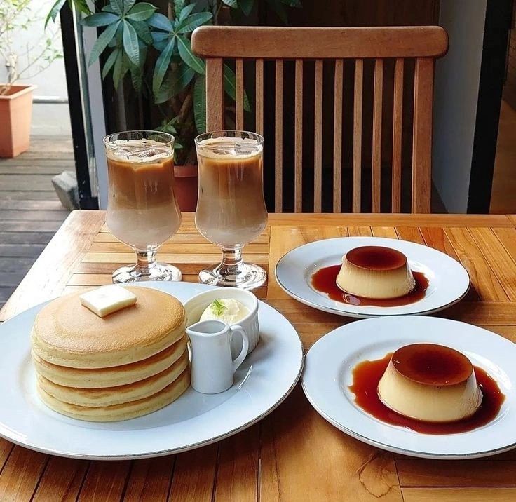 Iced coffee, pancakes with butter, and two plates of flan on a wooden table
