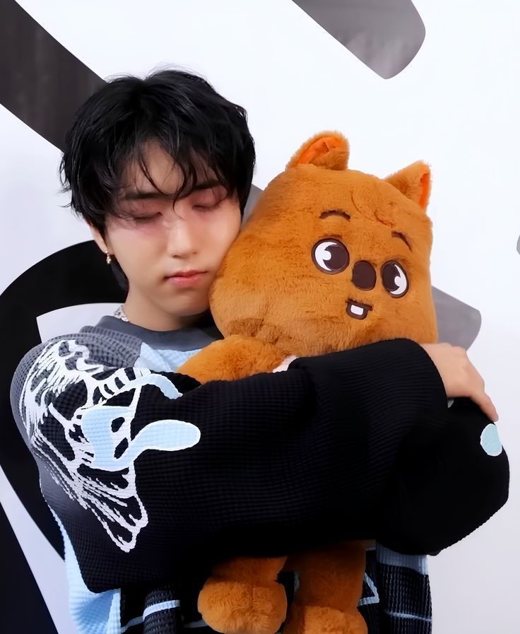 Young man with black hair hugging a brown stuffed animal plush toy against a white background